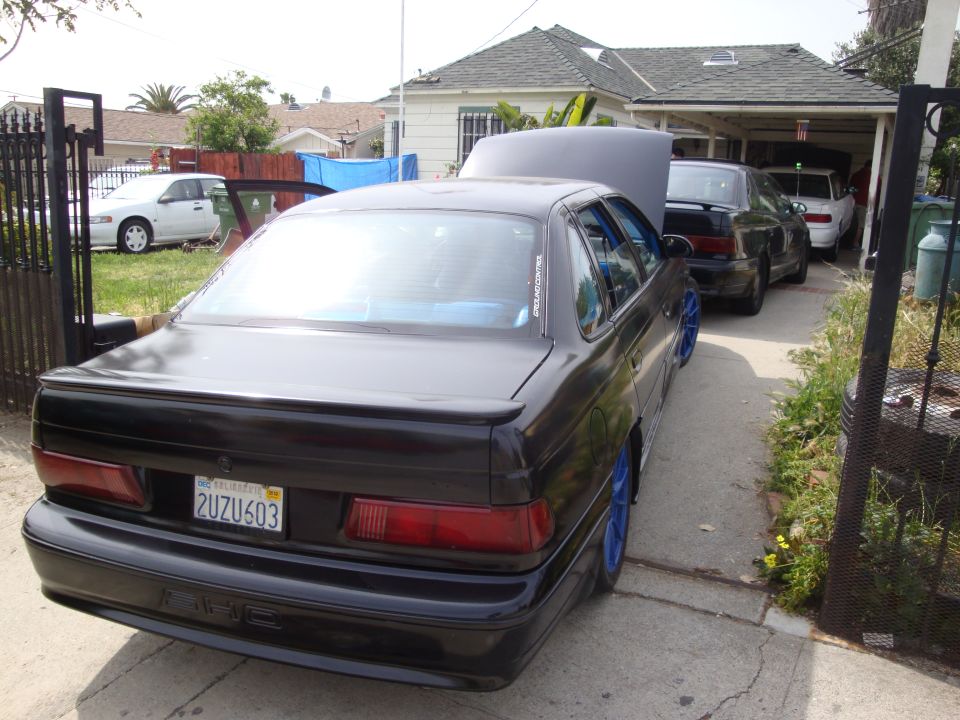 Black sedan with blue wheels and hood up, parked in driveway with two other cars. License plate 2UZU603