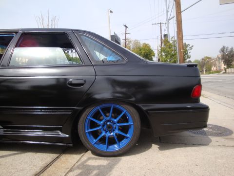 1995 Ford Taurus - A black sedan with custom blue wheels is parked on the side of the road.