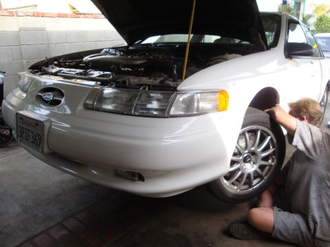 1995 Ford Taurus - A person works on the front wheel of a white Ford Taurus with its hood open.