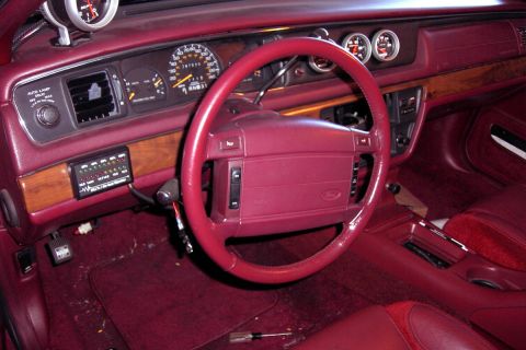 1992 Ford Crown Victoria - Burgundy interior of a Ford sedan shot from the driver's door, showing the red four-spoke airbag steering wheel, woodgrain dash trim, and a…