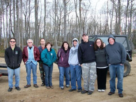 1986 Ford Bronco - A group of people stand in front of a dark SUV in a wooded area.
