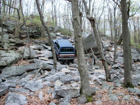 1986 Ford Bronco - Blue SUV navigates a challenging rocky trail through a wooded area.