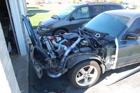 1985 Mercury Grand Marquis - The front end of a dark blue sedan is disassembled, revealing a modified engine and turbocharger, with a grey minivan in the background.