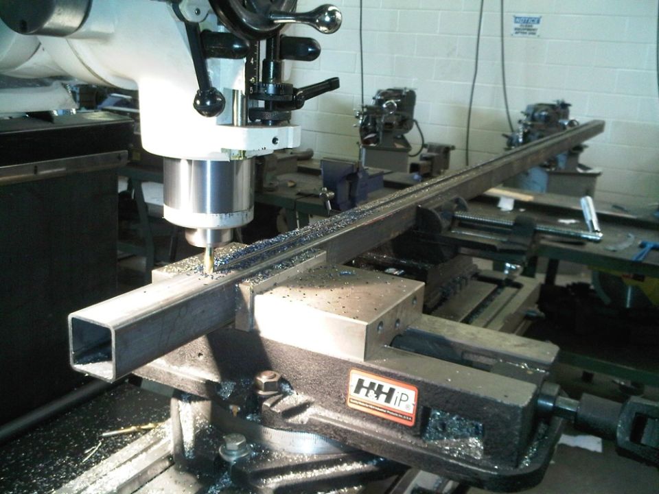 Milling machine cutting a square metal bar held in a vise, metal shavings visible