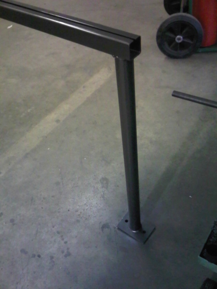 Metal railing post with a square top section and a round vertical support, bolted to a square base on a concrete floor