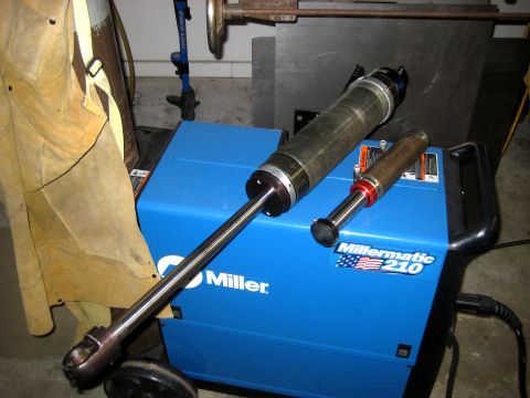 1996 Ford Bronco - Two hydraulic cylinders are placed on top of a blue Miller Millermatic 210 welding machine, with a leather apron visible to the left.