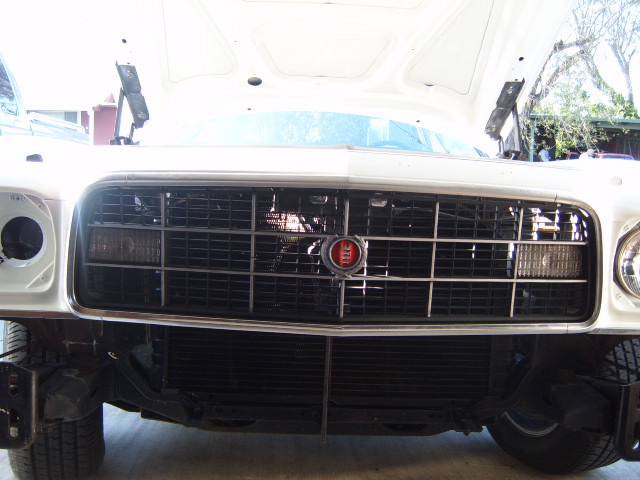 White car's front grille with a red emblem and horizontal bars, showing exposed radiator and tire