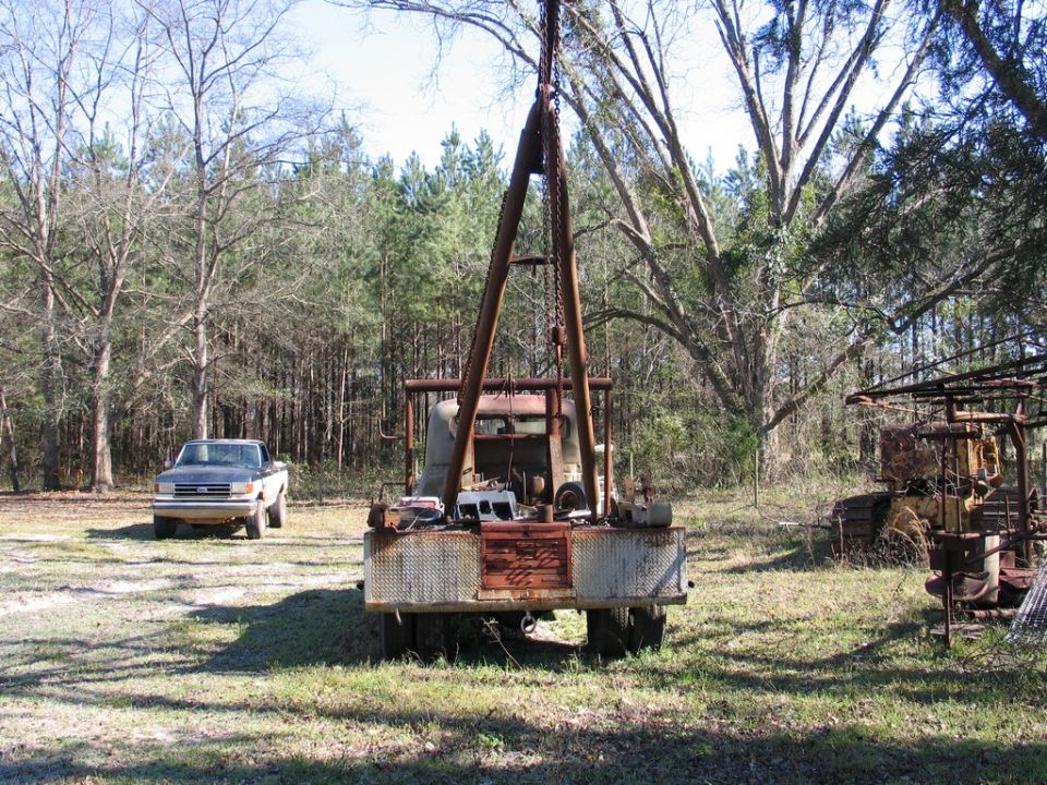 Rusty flatbed wrecker truck with boom and chain hoist parked in a field