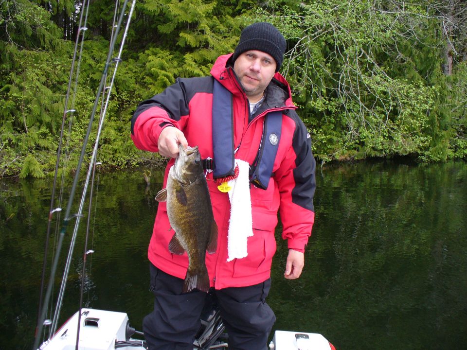 Angler in red jacket and life vest holding a smallmouth bass in a boat on a forested lake