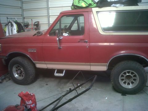 1989 Ford Bronco - This red Ford Bronco, likely from the late 1980s, is parked indoors next to lawn equipment.
