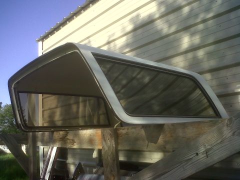1989 Ford Bronco - Truck cap with tinted windows, resting on wooden beams in front of a building.