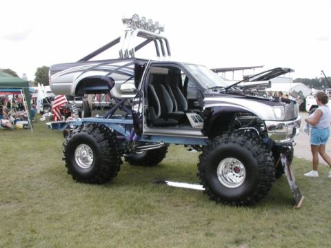 1994 Ford Bronco - This heavily modified pickup truck features massive tires and a lifted chassis, displayed outdoors with people and tents in the background.