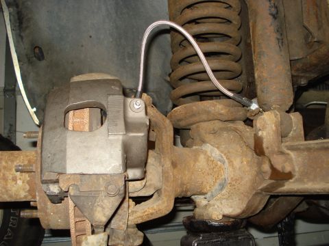 1979 Ford Bronco - Close-up under a truck showing a cast front brake caliper mounted at the knuckle, fed by a braided stainless steel flex brake line clamped…