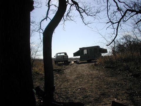 1979 Ford Bronco - A white lifted truck parked beside a set-up pop-up tent camper at a rough dirt campsite on a ridge, shot from below through bare winter tre…