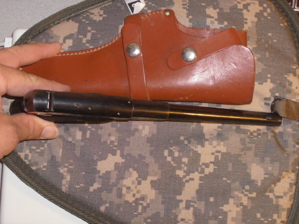 Hand holding a black and brass object near a brown leather holster on camo fabric