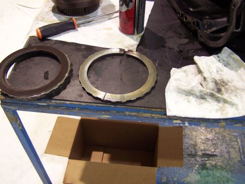 1973 Ford Ranchero - Transmission clutch pack and a damaged ring are laid out on a workbench next to a screwdriver and a can of spray.
