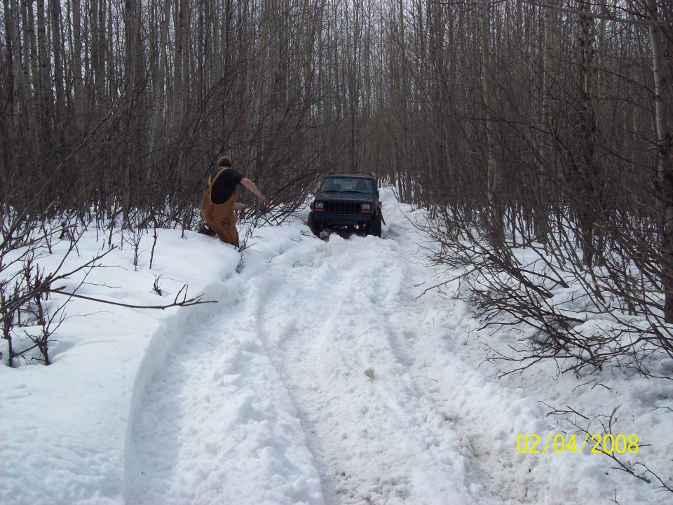 Jeep Cherokee stuck in snow on a wooded trail. Person in overalls is near the vehicle