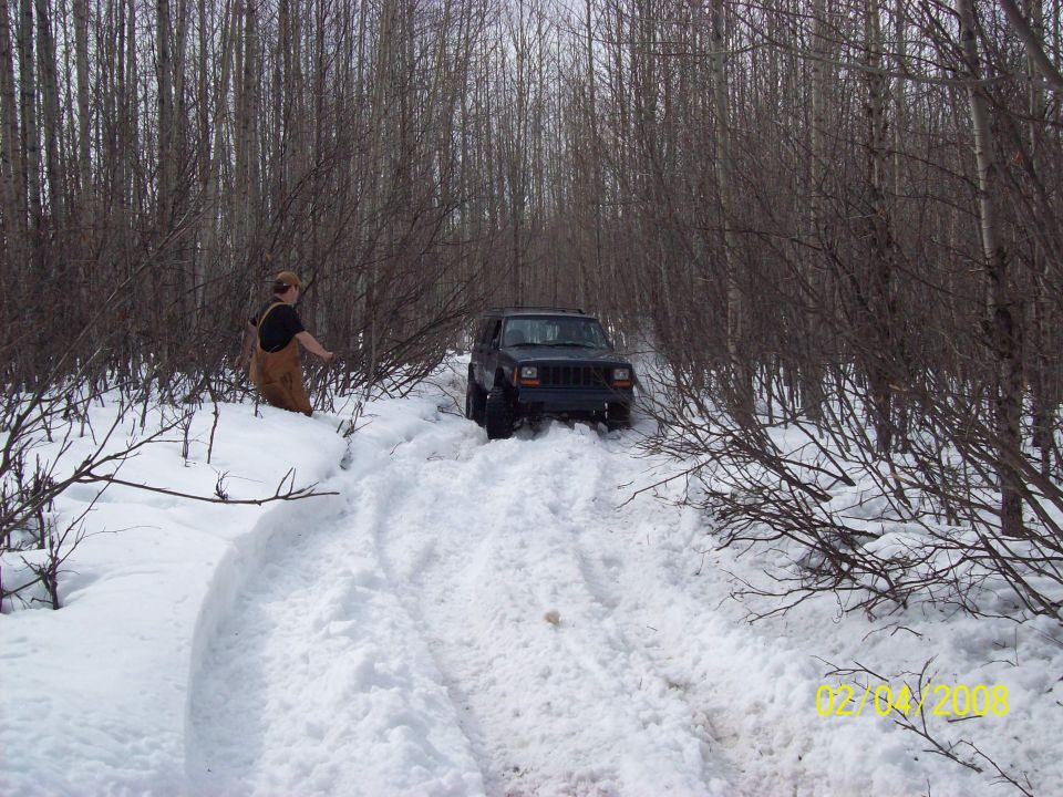 Jeep Cherokee XJ driving through deep snow in a wooded area. Person in overalls stands nearby