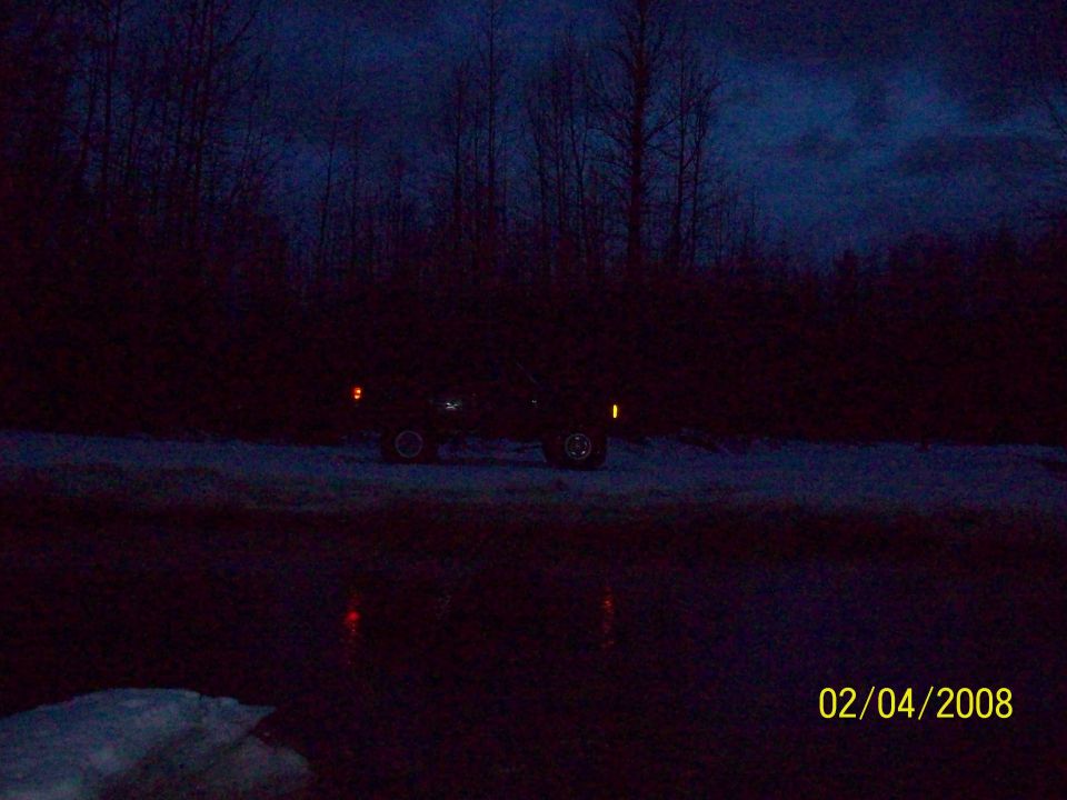 dark SUV parked on snowy ground near trees at dusk, headlights on