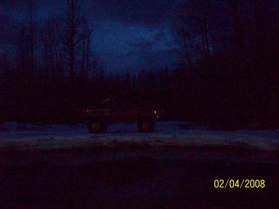Dark silhouette of a pickup truck with headlights on, parked in a snowy, wooded area at dusk. Watermark reads 02/04/2008 SUPERMOTORS.NET