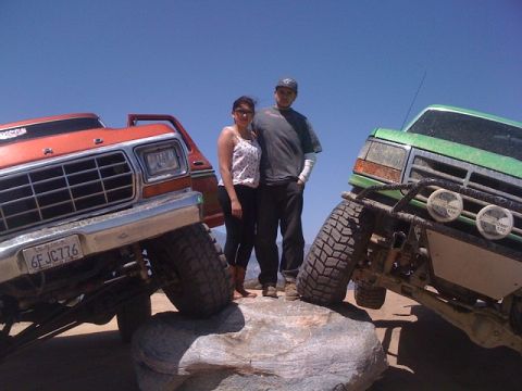 1978 Ford Bronco - Two people stand between a lifted orange truck and a green truck on a rocky terrain under a clear blue sky.