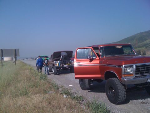 1978 Ford Bronco - An orange 1978 Ford Bronco is parked on the side of the road with its door open, while another vehicle is being worked on in the background.