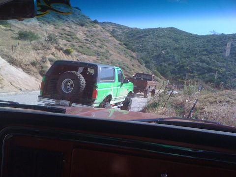 1978 Ford Bronco - Two trucks, a green Bronco and a brown pickup, drive on a dirt road through a hilly, vegetated area.