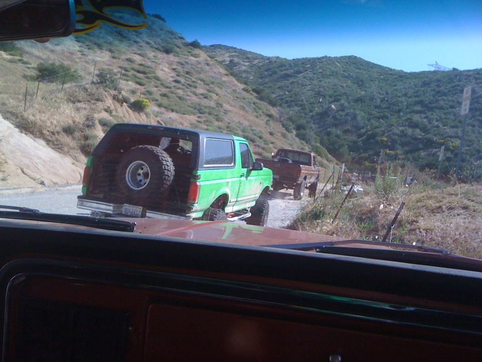 Green Ford Bronco with spare tire mounted on rear door, followed by brown Ford pickup truck on a dirt road