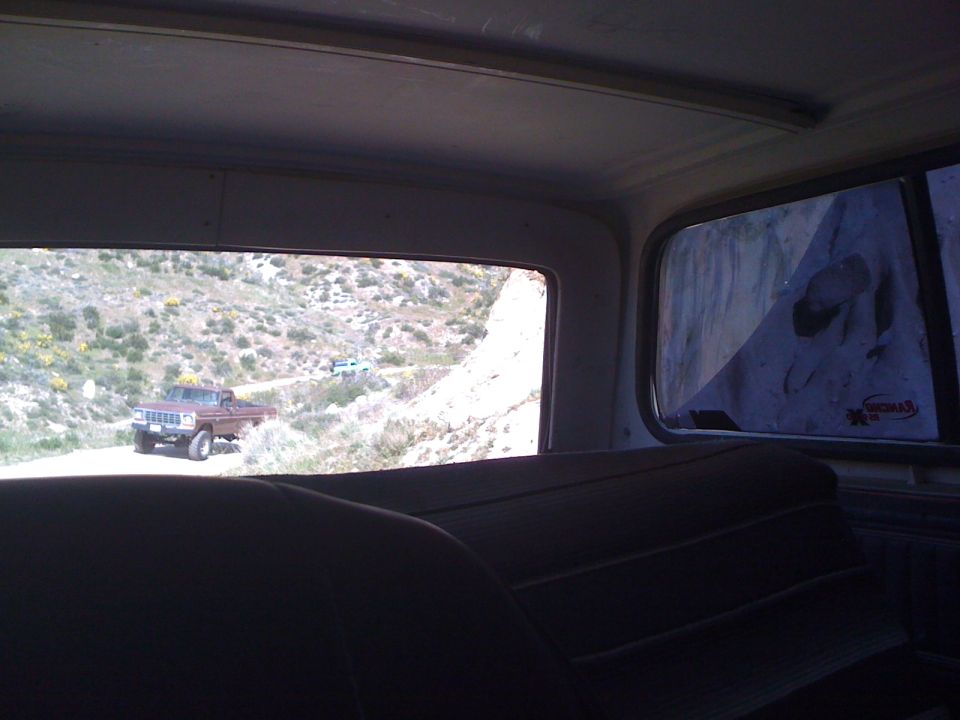 View from inside a vehicle looking out at a brown pickup truck on a dirt road in a hilly landscape. Another vehicle is visible further up the road