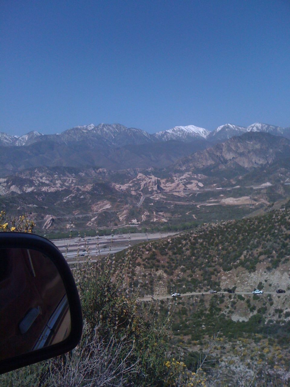 Side mirror view of a mountain road with several cars driving, snow-capped mountains in the distance