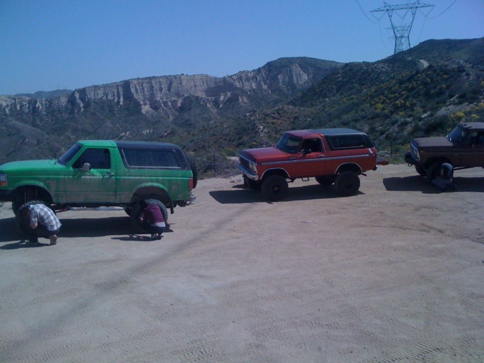 Three Ford Broncos parked on a dirt road, two green and red, with a brown one further back. People are working on the green Bronco