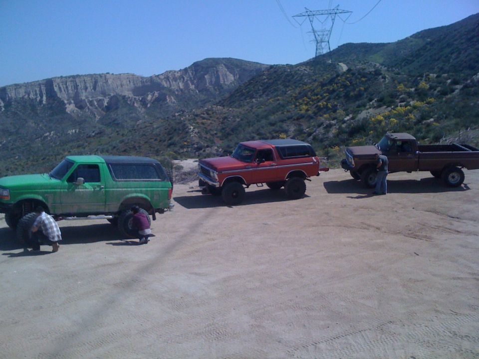 Three off-road vehicles parked on a dirt lot with mountains in the background. Two people are working on the green Bronco