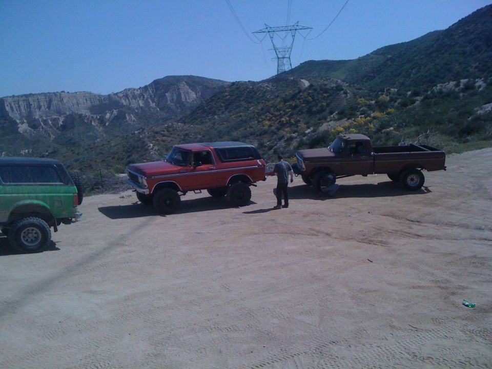 Red Bronco, green Bronco, and brown pickup truck parked on dirt road with hills and power lines in background