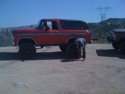 1978 Ford Bronco - A person works on a tire of a red 1978 Ford Bronco parked next to another vehicle outdoors.
