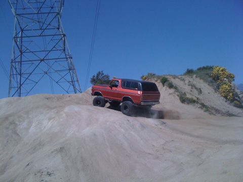 1978 Ford Bronco - This orange Ford Bronco is kicking up dust as it drives up a sandy hill under a clear blue sky.