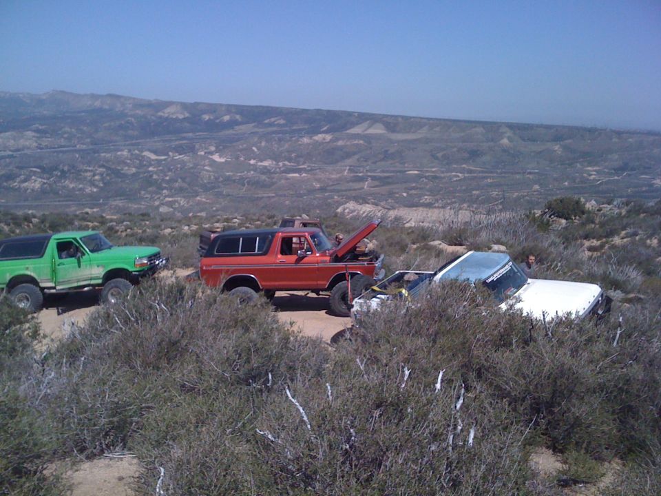 Three off-road vehicles parked on a dirt path in a dry, hilly landscape. One is green, one is red with its hood open, and a white truck is partially obscured by bushes
