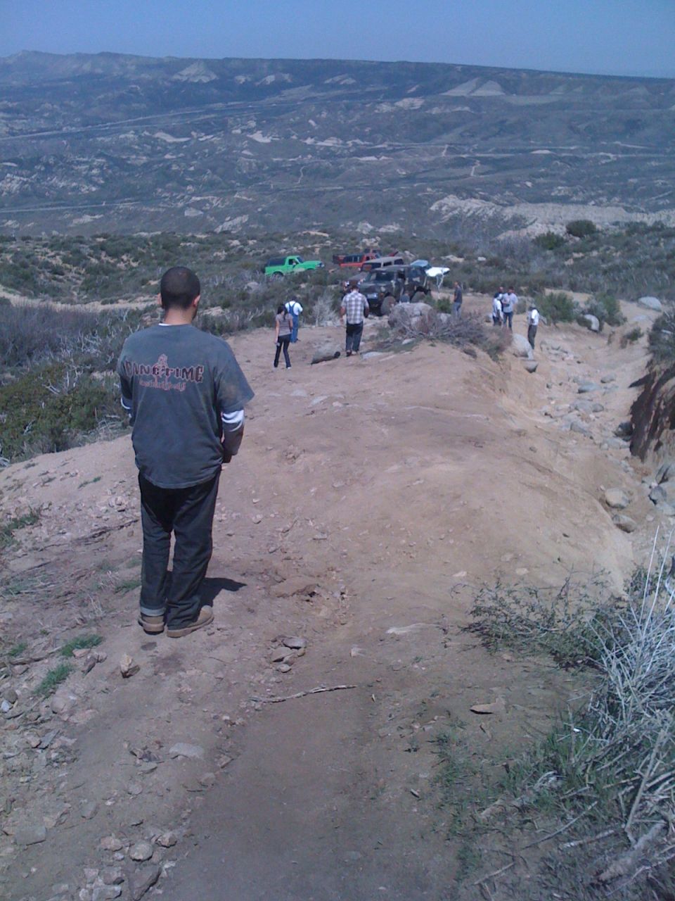 Several off-road vehicles parked on a dirt trail with people standing around, overlooking a valley