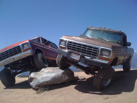 1978 Ford Bronco - Two lifted Ford trucks, a brown F-series and a red Bronco, navigate rocky terrain under a clear blue sky.