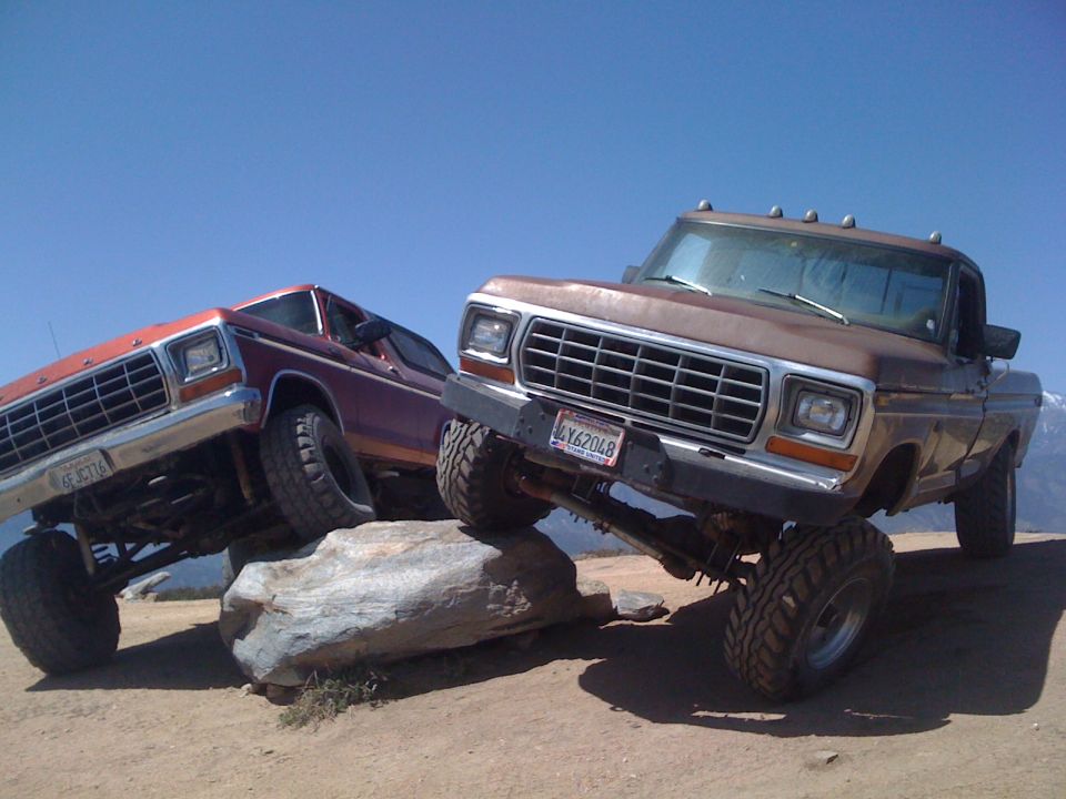Two lifted Ford trucks, a brown F-series and a red Bronco, are off-roading over rocks. The brown truck has license plate LY62048