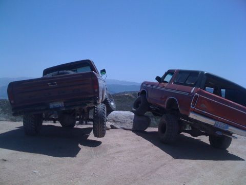1978 Ford Bronco - Two lifted Ford trucks navigate a rocky off-road trail under a clear blue sky.