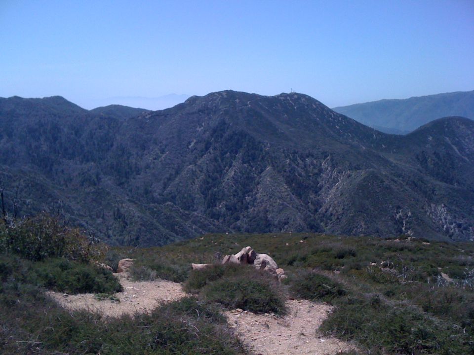 Mountain range with trees and a clear blue sky. A dirt path is visible in the foreground