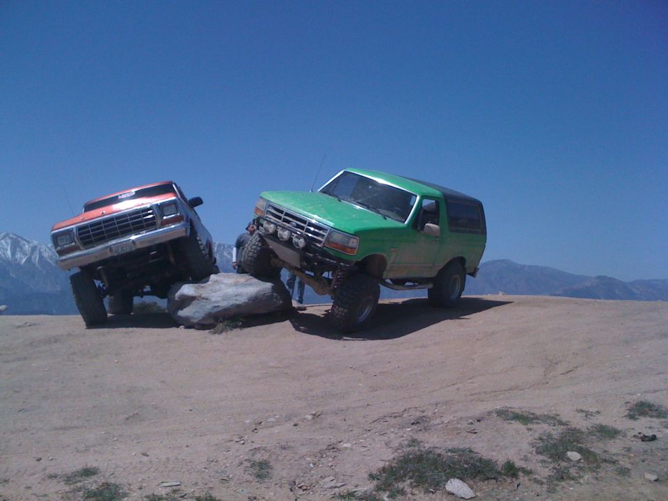 Green 1987 Ford Bronco climbing rocks, orange Ford truck behind