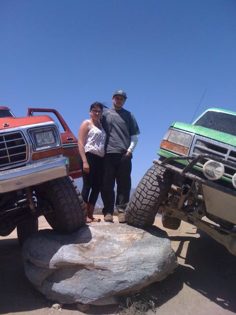 1978 Ford Bronco - A couple stands on a large rock between two heavily modified pickup trucks under a clear blue sky.