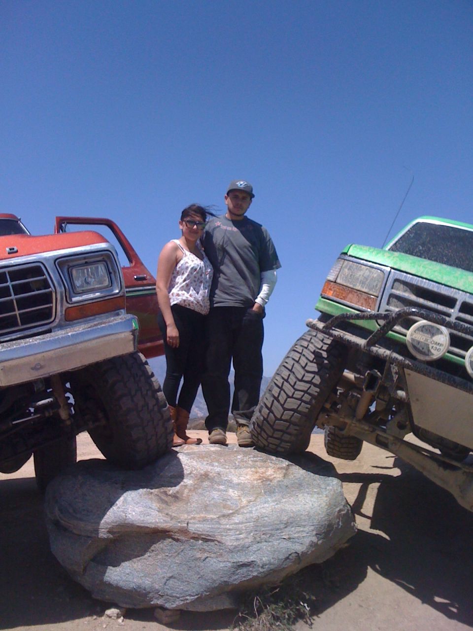 Couple posing between a red and green lifted pickup truck on a rock