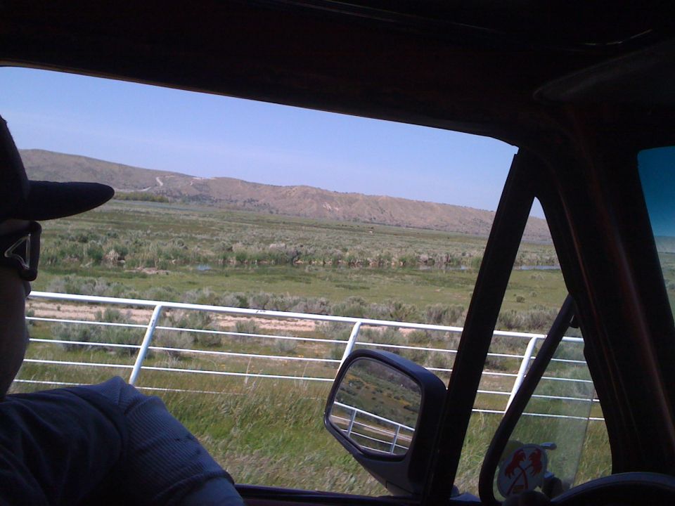 Interior view of a vehicle with a person wearing a hat, looking out at a rural landscape with mountains and a fence