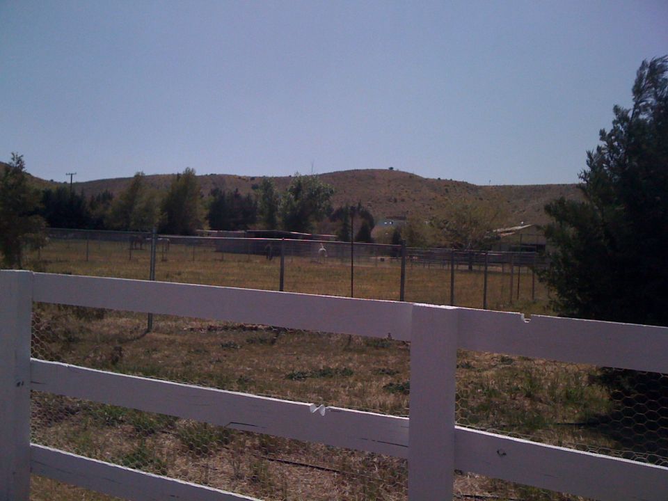 White wooden fence in foreground, dry grass field, chain-link fence, trees, and rolling hills under a clear blue sky. Supermotors logo visible