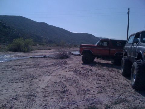 1978 Ford Bronco - Two lifted off-road trucks, including a 1978 Ford Bronco, are parked on a dirt road in a scenic, hilly area.