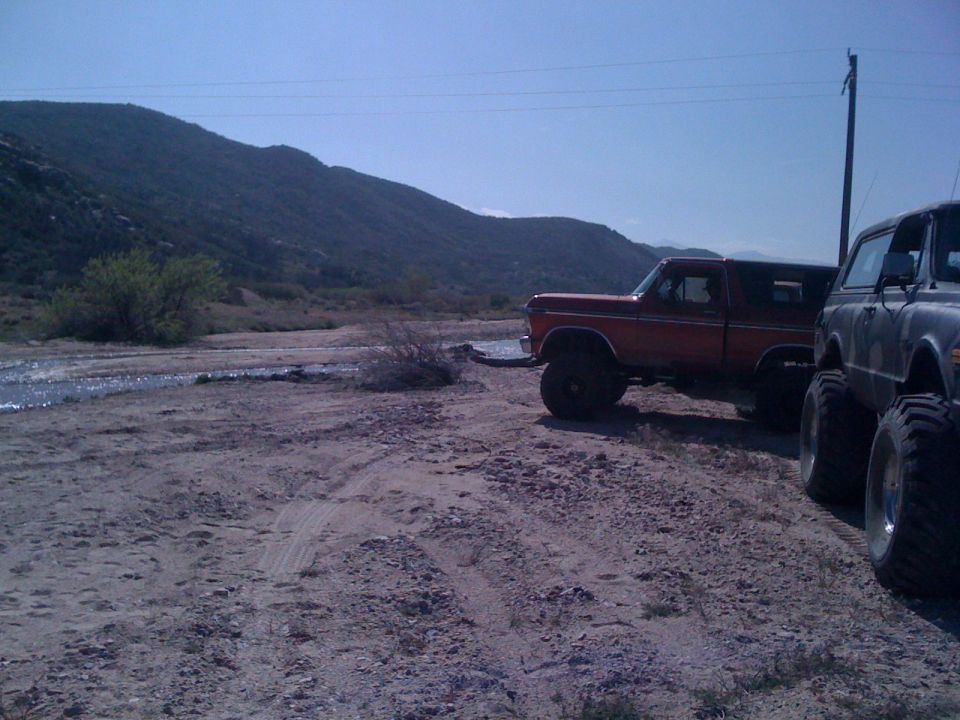 1978 Ford Bronco and another lifted truck parked on a dirt road near a stream with hills in the background