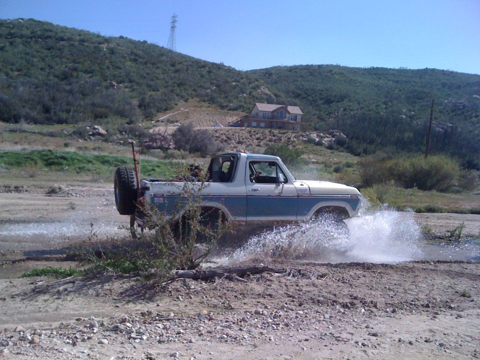 Ford Bronco driving through a shallow stream, kicking up water. House on hill in background