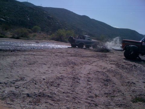 1978 Ford Bronco - Two trucks navigate a dusty, off-road terrain with a body of water in the background.